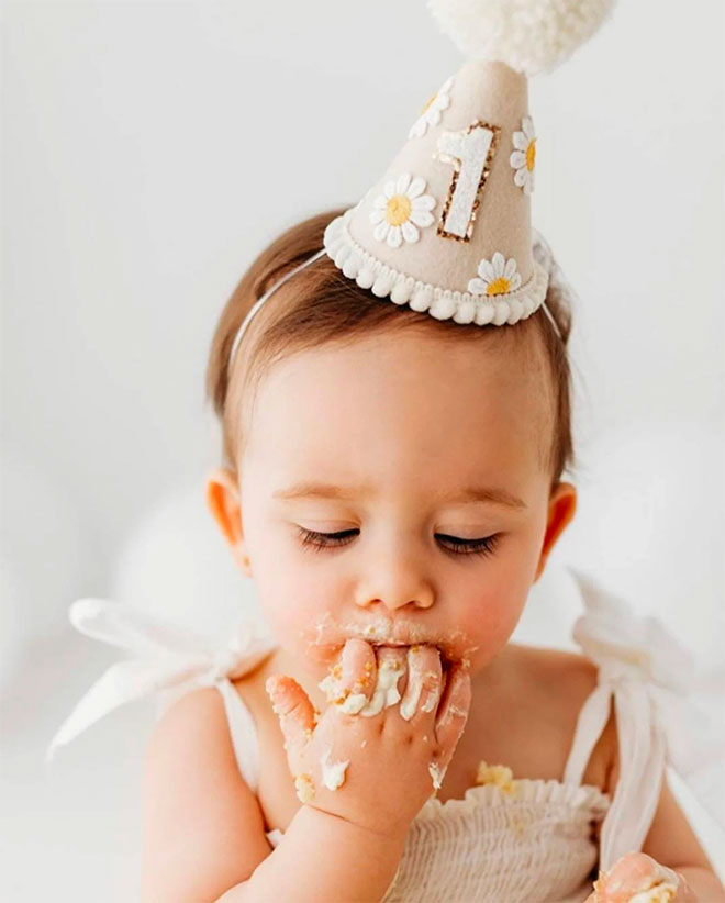One year old girl eating cake of her fingers in a Daisy birthday hat from Our Little Dear