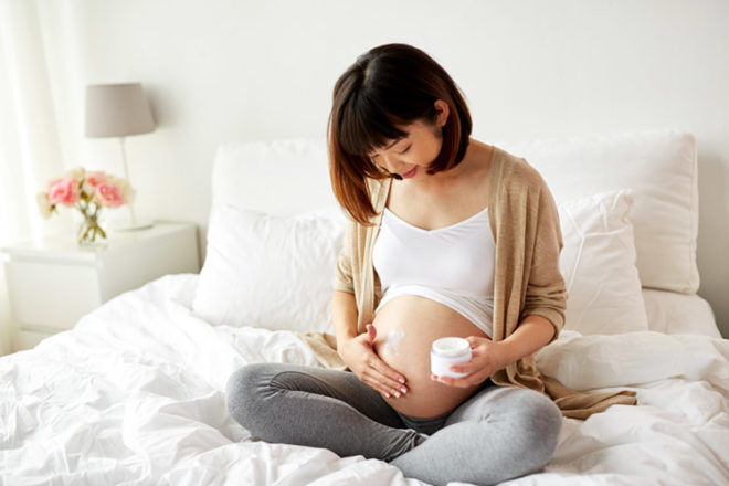 Pregnant woman showing herself applying a white bottle of stretch mark cream to her belly