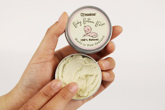Haakaa Baby Bottom Balm tin being held in ladies' hand. The tin is open showing the colour and consistency of the cream inside. 