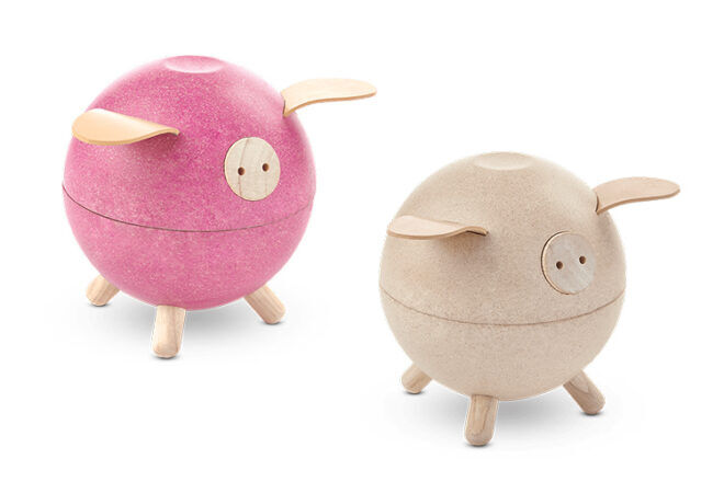 Plan Toys showing wooden Piggy Banks in natural and pink shades with three legs