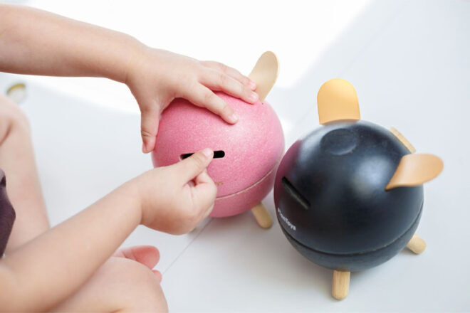 Plan Toys Piggy Banks showing a young child putting a coin into the pink money box