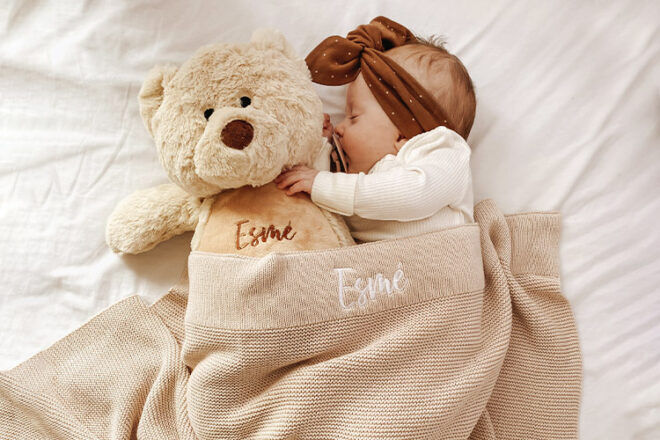 Newborn baby sleeping under Little Poppet Store embroidered baby name blanket, showing name on border on the beige colourway, and matching personalised teddy bear. 