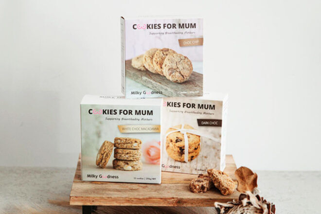 Freshly baked Milky Goodness Cookies for Mum is shown, with three varieties of breastfeeding support cookies in their respective boxes positioned behind them. Each box clearly displays the flavor of the cookies along with the key ingredients. One of the cookies is broken in half, showing its texture and visible ingredients such as oats, flaxseed, and brewer's yeast.
