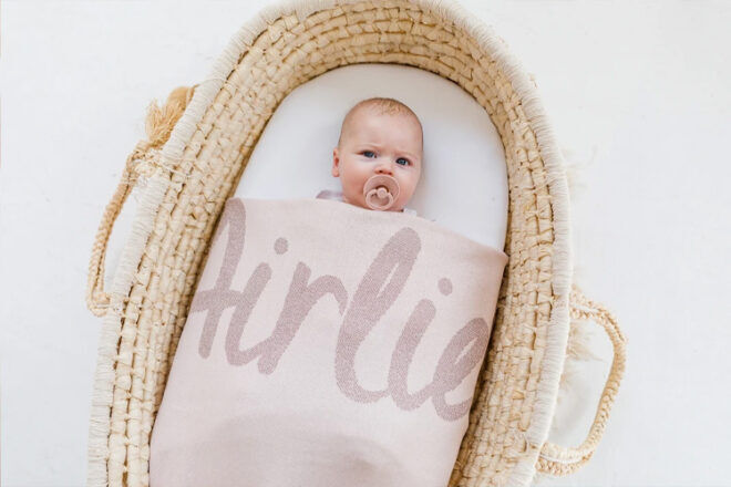 Birdseye view of a baby girl in a Moses basket, tucked into a Namely Co custom baby blanket, showing the large name Airlie in a script font in the Rose Pink and Lurex Rose Gold colourway.