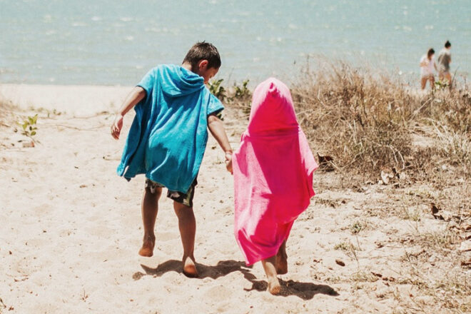 Back view of young boy and girl wearing Wovii Kids toddler bathrobes at the beach, showing hood off and hood on, as well as two different colours in blue and pink. 