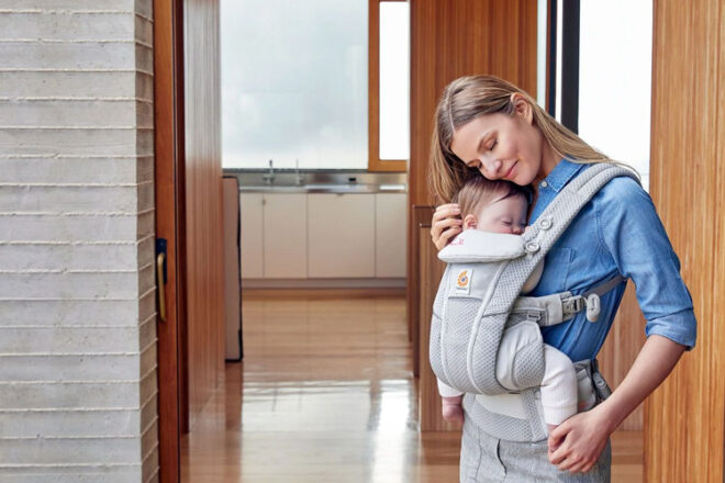 Woman wearing sleeping baby in Ergobaby Omni Breeze Carrier, showing mesh lining, adjustable straps and that it's a gift idea for new parents.
