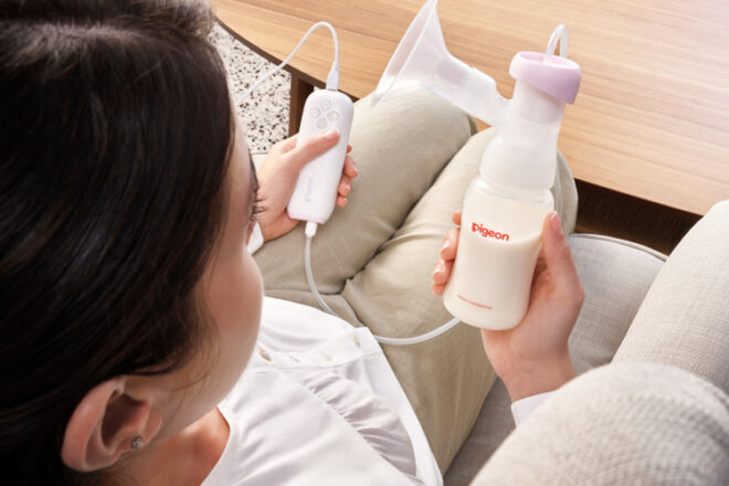 Pigeon GoMini Breast Pump being used by a mother on the couch. She is holding the bottle of milk and the small controller in the other hand. 