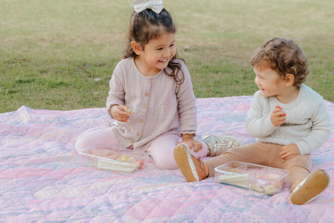 Two toddlers sitting on an Emro Designs park blanket showing pastel print, practical, quilted surface and size in comparison to the children.