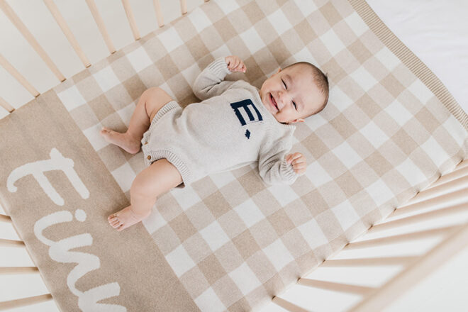 Birdseye view of baby in cot lying on top of a Namely Co baby name blanket, showing large name Finn in Camel Gingham design.