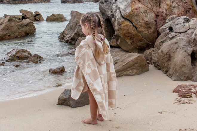 Back view of young girl at the beach wearing Calf & Crew terry kid hooded towel, showing unique checkered design, hood and length of towel in comparison to the child.