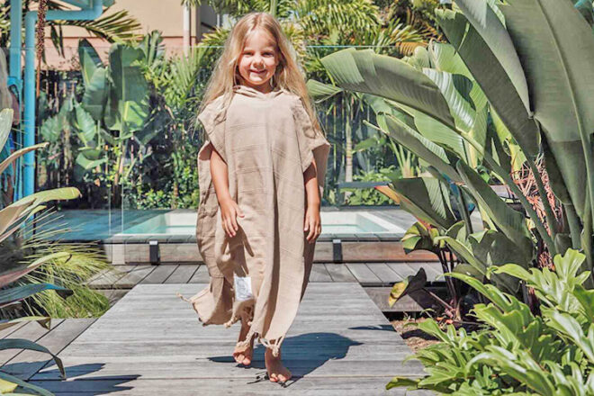 Front view of young girl wearing Chasing Sundays Turkish hooded poncho towel, showing use in a pool setting as well as the long length of the towel for coverage, fringe trim and beige colourway.