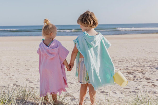 Back view of two young children at the beach wearing Coast Kids beach towels with hood, showing hood, fringe edge and two different colours, pink and blue.