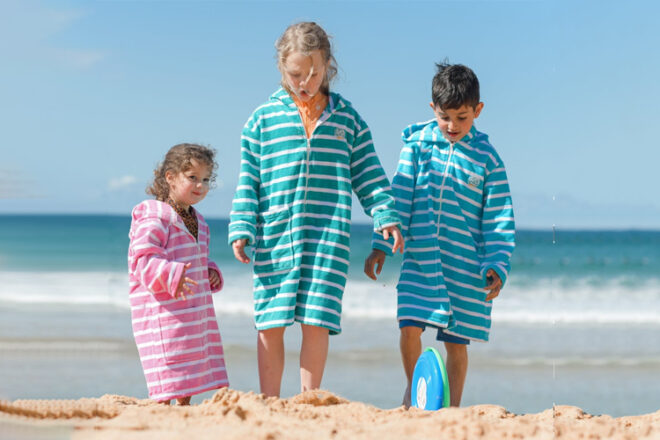 Front view of three children at the beach wearing Rad Kids Hooded Poncho Towels showing different sizes for different ages, all three colours and how the towels can be zipped up for coverage. 