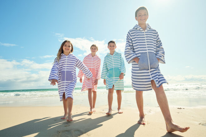 Four smiling children at the beach wearing Sammimis Turkish hooded poncho towels showing them zipped up for coverage, all four colours, as well as large front pockets. 