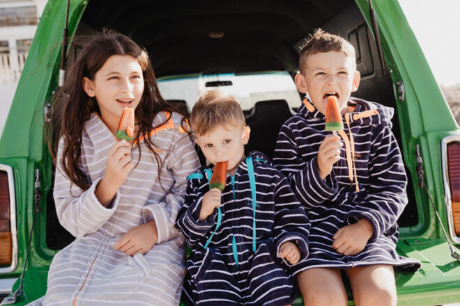 Three children sitting in the back of a car wearing Swoodi surf poncho towels, showing different colourways, that the towels come in various sizes for different ages, and that they are full coverage. 