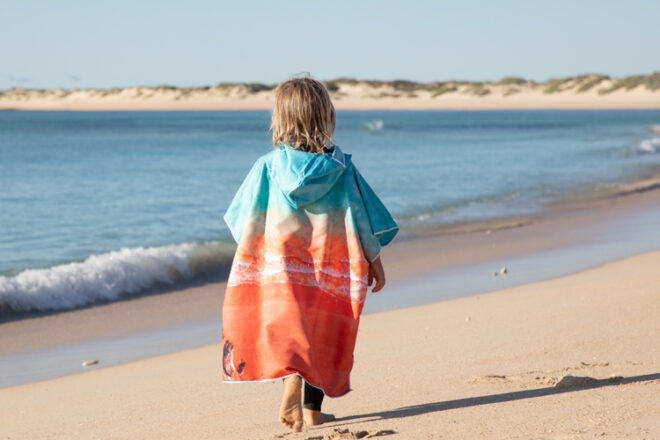Back view of young child walking along the beach wearing Will & Wind microfiber baby hooded towel, showing unique beach print.