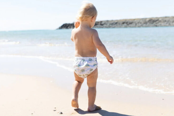 Back view of toddler wearing Bare & Boho reusable swim nappy, showing beach setting and pam leaf print.