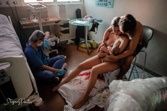 Postpartum photo of couple cuddling new born and exhausted midwife sitting on floor