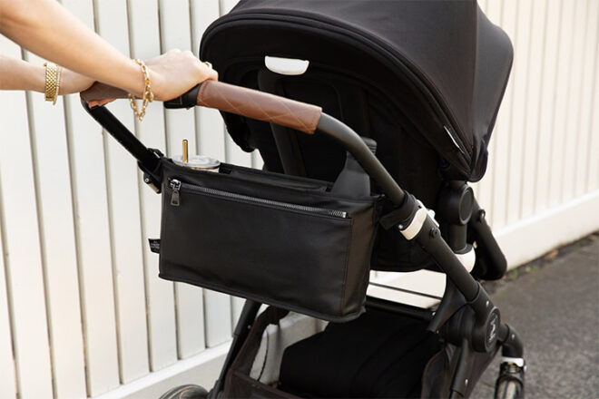 The Nappy Society Vegan Leather Pram Caddy attached to a pram showing a mum pushing the stroller whilst a drink is in the bag. 