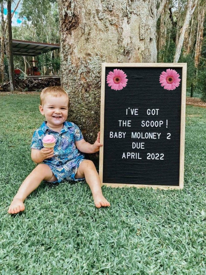 A toddler sits next to a sign saying “i’ve got the scoop, baby 2 due April 2022'
