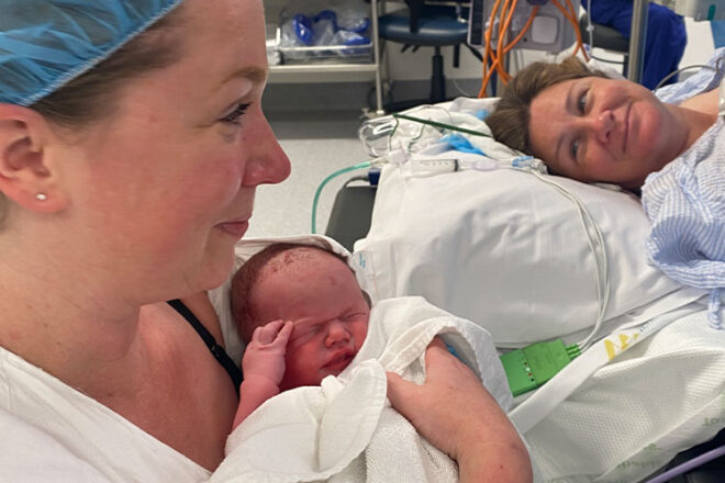 Danni holds her baby in the delivery room with her surrogate sister in law smiling on the bed