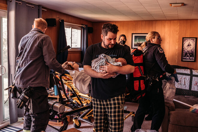 A man holding his newborn baby surrounded by medical staff and his wife on a stretcher