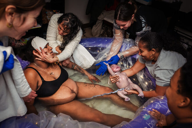 A woman who has just given birth in a paddle pool with her baby still connected via umbilical cord surrounded by women