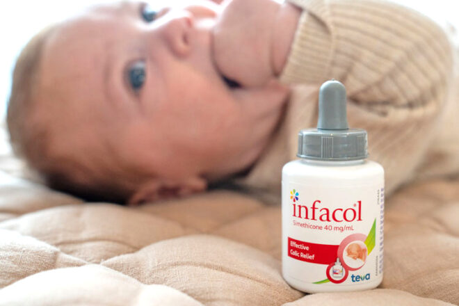 Baby on a bed with hand in mouth lying next to a bottle of Infacol 