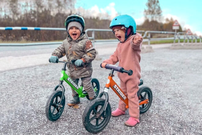 two young children sitting on a strider balance bike one green and one orange