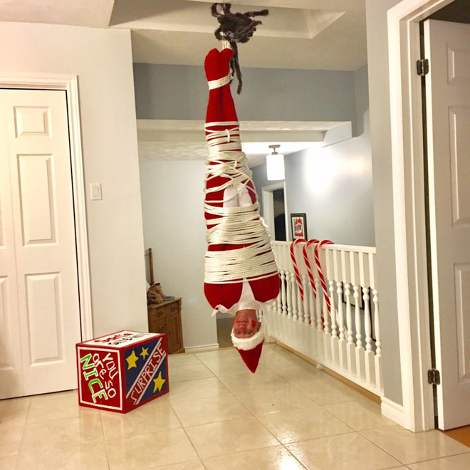 Dad dresses as elf on the shelf hanging upside down and tied up like mistletoe