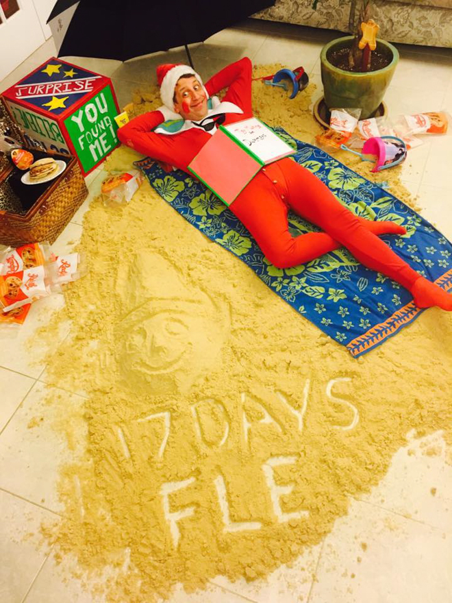 Dad dresses as elf on the shelf laying on a towel with sand all around him pretending to be on the beach