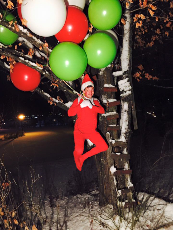 Dad dresses as elf on the shelf whilst pretending to be floating by holding onto a bunch of balloons stuck in a tree