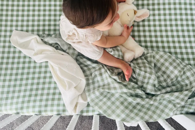 Baby sitting in a cot holding a teddy and comfort blanket