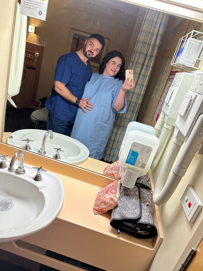 Tenae in a blue hospital gown and husband taking a selfie in the bathroom mirror