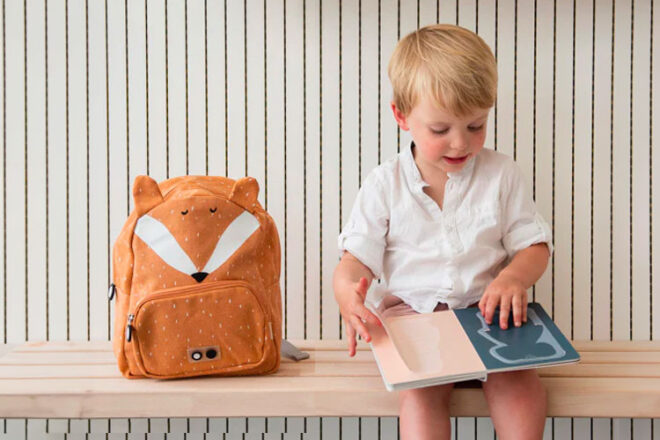 Little boy sitting on a bench reading nect to the TRIXIE Fox Backpack