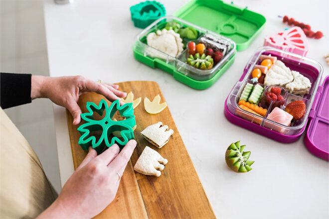 Lunch Punch sandwich cutters in dinosaur feet shapes next to packed lunch showing a mum cutting the food. 