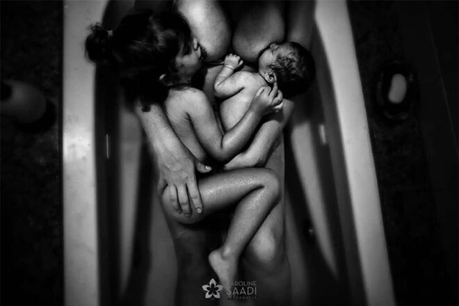 A mother laying in a bathtub breastfeeding her toddler and newborn baby