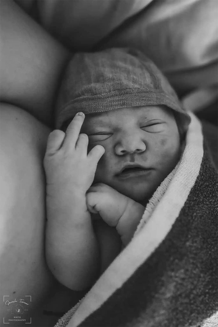A newborn baby swaddled in the arms of a person holding up their middle finger